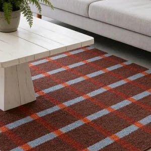 Può includere: Un tappeto rettangolare con un motivo a quadri nei toni del marrone, arancione e azzurro. Il tappeto è posizionato sul pavimento sotto un tavolino bianco e un divano grigio. Il design del tappeto presenta linee che si intersecano.