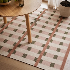 Puede incluir: Una alfombra rectangular con un patrón de cuadrícula de bloques en color crema, rosa empolvado y verde oliva. El diseño incluye líneas verticales y horizontales prominentes en color terracota, creando un aspecto tejido y estructurado.