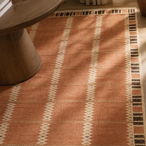 May include: A rectangular area rug featuring a terracotta orange base colour with vertical rows of cream-coloured diamond patterns. The border consists of alternating dark brown and orange rectangular blocks, finished with a short fringe at the edge.