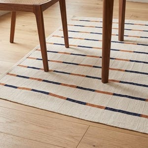 May include: A rectangular rug with horizontal stripes in alternating navy blue and rust-orange on a cream background. The rug is partially under the legs of a wooden chair and table.