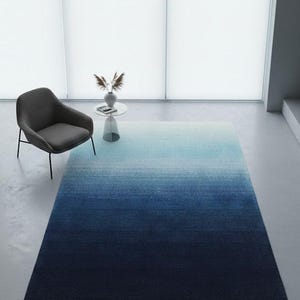 Pode incluir: Um tapete retangular com um design gradiente, que transita de azul claro no topo para azul marinho escuro na parte inferior. Uma poltrona cinza e uma pequena mesa com um vaso e objetos decorativos estão no fundo.