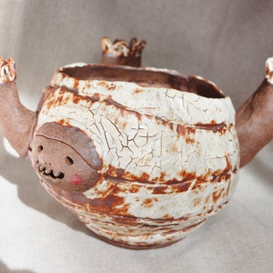 Puede incluir: Un macetero de cerámica blanca con forma de momia con detalles marrones y una cara sonriente. El macetero tiene dos brazos y un pico.