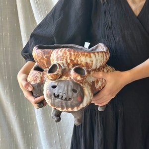 Puede incluir: Una maceta de cerámica marrón y blanca con una cara caprichosa y dos pequeños agujeros en la parte superior. La maceta tiene una superficie texturizada y parece estar hecha a mano.