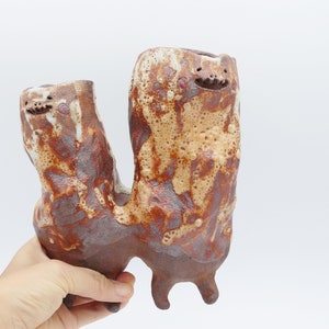 May include: A ceramic sculpture of two figures with brown and white glaze. The figures have a whimsical, abstract design and appear to be connected.