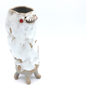 Puede incluir: Un jarrón de cerámica hecho a mano con una forma orgánica única. El jarrón es principalmente blanco con un esmalte texturizado y goteado, y presenta un diseño similar a una cara con acentos rojos. Se apoya sobre tres pequeños pies.