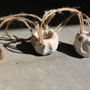 May include: Two small white ceramic planters with rough textures are suspended by twine. The planters are hanging from a single piece of twine, which is looped around the top of each planter.