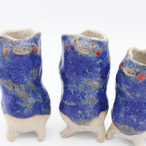 Puede incluir: Tres jarrones de cerámica azul y blanco con detalles rojos y un diseño caprichoso de estilo de dibujos animados. Cada jarrón tiene tres patas y una superficie texturizada.
