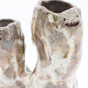 May include: A handcrafted ceramic vase with a unique, organic shape. The vase features two openings and is decorated with a mottled, earthy color palette of brown, white, and hints of red. The design includes whimsical, smiling faces.