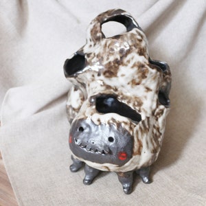 May include: A ceramic sculpture of a creature with a brown and white speckled body, black eyes, and red cheeks. The creature has four legs and a hole in the top of its head.