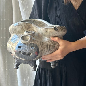 Puede incluir: Una maceta de cerámica con un diseño caprichoso y texturado. Presenta una cara con una amplia sonrisa y dos pequeños agujeros en la parte superior. La maceta está pintada en tonos grises, blancos y marrones.