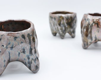 May include: Three small, ceramic pots with a textured, speckled glaze in shades of brown, black, and white. The pots have four legs each and are arranged on a white surface.