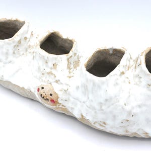 Może przedstawiać: Ręcznie robiona ceramiczna donica z czterema otworami. Donica jest podłużna, z teksturowanym białym szkliwem na beżowej podstawie. Mała, fantazyjna twarz z czerwonymi akcentami jest widoczna z boku. Idealna do sukulentów.