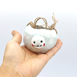 Puede incluir: Una pequeña maceta de cerámica blanca con diseño de rostro, con detalles rojos en las mejillas y la boca. La maceta tiene un lavado azul claro y se sostiene en una mano. Cuerda natural atada en la parte superior.
