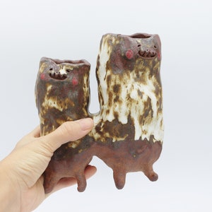 May include: Two ceramic planters shaped like cartoon characters with brown and white glaze. The planters have red painted details on their faces and stand on three legs.