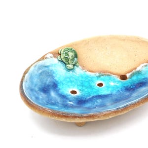 Könnte beinhalten: Keramik-Seifenschale mit Strand- und Ozean-Design. Die Schale hat einen hellbraunen Sandbereich, türkisfarbene Wellen und eine kleine grüne Schildkröte. Die Schale hat drei Abflusslöcher und wird von drei kleinen Füßen getragen.