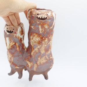 May include: A ceramic vase with a whimsical design featuring two brown and white figures with smiling faces. The vase has a textured surface and a unique, abstract shape.