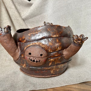 Puede incluir: Un cuenco de cerámica hecho a mano con un exterior marrón texturizado. El cuenco presenta una cara de dibujos animados con una boca sonriente y brazos extendidos. El cuenco es un objeto decorativo único. Medidas en centímetros.