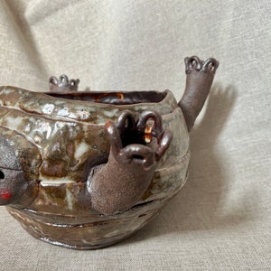 Puede incluir: Un macetero de cerámica marrón y blanco con forma de tortuga con los brazos levantados y una cara. El macetero tiene un acabado esmaltado y una superficie texturizada.