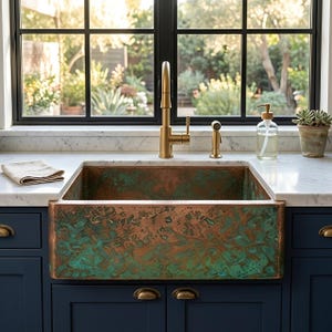 May include: A farmhouse sink with a hammered copper and green patina finish, set into a white marble countertop above dark blue cabinets. A tall brass faucet, a side sprayer, a clear glass soap dispenser, and a small potted succulent are present.