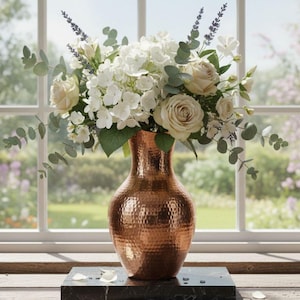 Könnte beinhalten: Eine kupferfarbene Vase mit Hammerschlag-Textur enthält ein Arrangement aus weißen Rosen, Hortensien und Grün. Die Vase steht auf einem dunklen quadratischen Sockel vor einem Fenster mit Gartenblick.