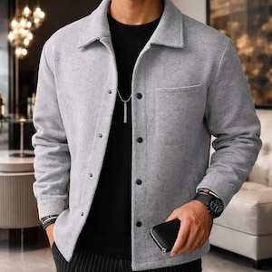 Könnte beinhalten: Hellgraue Button-up-Jacke mit Kragen und einer Brusttasche. Die Jacke hat schwarze Knöpfe und wird über einem schwarzen Hemd getragen. Eine schwarze Uhr und ein Armband sind am Handgelenk zu sehen. Eine schwarze Geldbörse wird in der Hand gehalten.