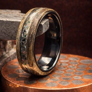 May include: A close-up of a ring with a black interior and a wooden exterior. The wooden exterior has a mix of light and dark brown tones with a section of colorful stone inlays. The ring is displayed on a copper-colored surface.