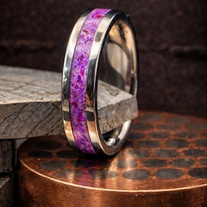 May include: A silver ring with a vibrant purple stone inlay. The ring features a polished silver band with a central channel filled with a textured, crushed stone material in shades of purple and pink.