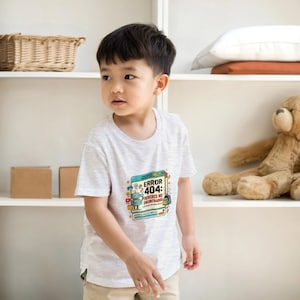 Könnte beinhalten: Ein hellgraues kurzärmeliges T-Shirt mit einem farbenfrohen Grafikdruck auf der Brust. Das Design zeigt eine kleine Roboter-Illustration und den Text "¡OOPS! ERROR 404: DEBERES NO ENCONTRADOS" sowie verschiedene kleine Symbole wie einen Fußball.