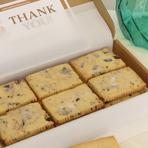 May include: A cardboard box containing rectangular shortbread biscuits with visible dark chocolate chips and a light dusting of sugar. A white paper insert inside the box features the text THANK YOU and the website address www.CakeCrumbCorner.com.