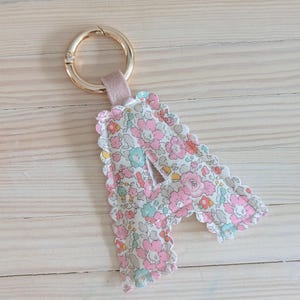 May include: A floral fabric letter "A" keychain with a gold-colored clasp and a pink suede strap. The letter is filled with a pink, white, and blue floral pattern and has a scalloped edge.