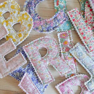 May include: A collection of fabric letters in various floral patterns, including yellow, pink, and blue, with white scalloped edges. The letters are arranged on a light wooden surface, creating a decorative display.