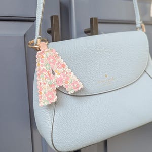 May include: A light blue leather crossbody bag with a matching strap and a floral print charm. The charm is in the shape of the letter "A" and is attached to the bag's strap with a gold ring.