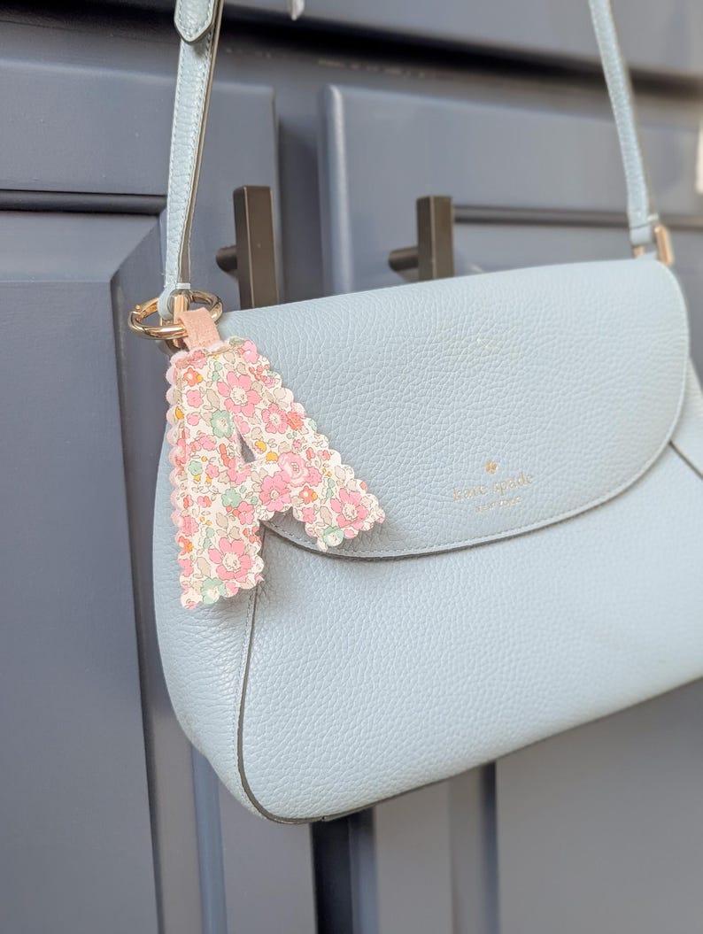 May include: A light blue leather crossbody bag with a matching strap and a floral print charm. The charm is in the shape of the letter "A" and is attached to the bag's strap with a gold ring.