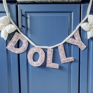 May include: A banner spelling "DOLLY" in pink glitter letters, strung on a white ribbon. The banner is adorned with two white fabric bows, one on each end. The banner is hanging against a blue cabinet.