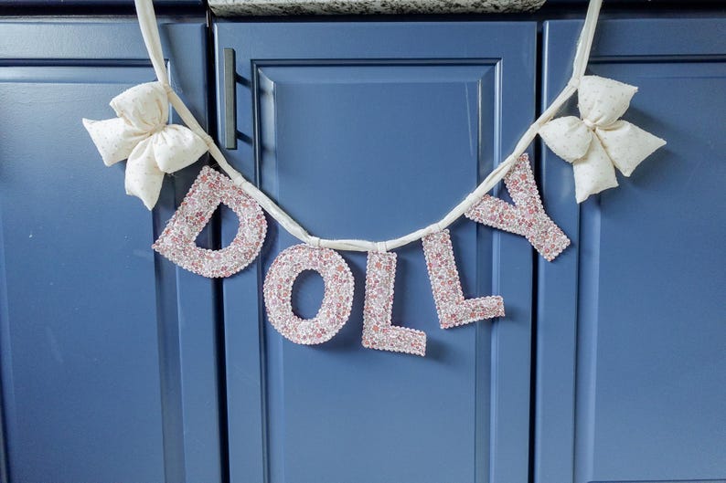 May include: A banner spelling "DOLLY" in pink glitter letters, strung on a white ribbon. The banner is adorned with two white fabric bows, one on each end. The banner is hanging against a blue cabinet.