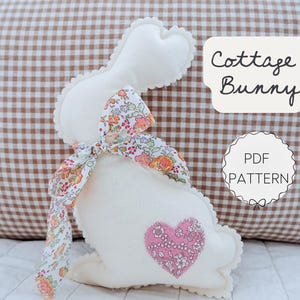 May include: A white stuffed bunny with a floral bow and a pink heart applique. The bunny has a scalloped edge and the text "Cottage Bunny" and "PDF Pattern" are visible.