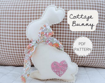 Puede incluir: Un conejito de peluche blanco con un lazo floral y un corazón rosa aplicado. El conejito tiene un borde festoneado. El texto "Cottage Bunny" y "PDF Pattern" son visibles.