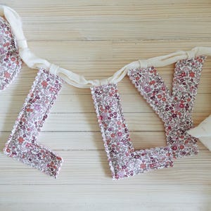 May include: A floral fabric banner spelling out the word "LOVE". The letters are made of a white and floral fabric with a scalloped edge. The banner is connected by a white ribbon.