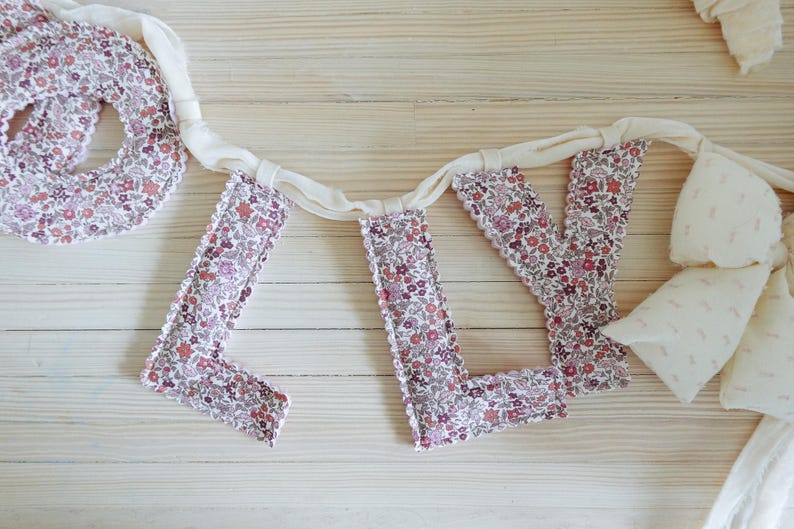 May include: A floral fabric banner spelling out the word "LOVE". The letters are made of a white and floral fabric with a scalloped edge. The banner is connected by a white ribbon.