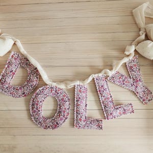 May include: A fabric banner spelling "DOLLY" in purple floral print letters. The letters are connected by a white ribbon and adorned with cream-colored bows at each end. The banner is displayed on a light wood surface.