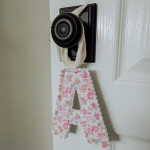 May include: A fabric letter "A" with a floral pattern in pink and white, trimmed with white lace, hangs from a door handle. The door handle is black and the door is white.