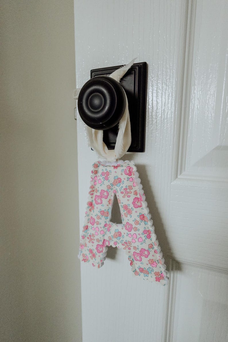 May include: A fabric letter "A" with a floral pattern in pink and white, trimmed with white lace, hangs from a door handle. The door handle is black and the door is white.