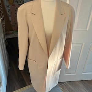 May include: A long-length blazer in a solid light beige color. This garment features a notched lapel collar, long sleeves with structured shoulders, and two large patch pockets positioned on the lower front section of the coat.