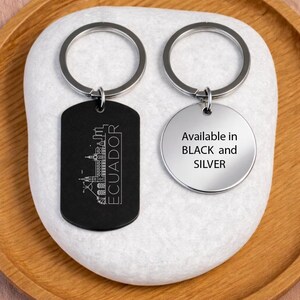 May include: Two keyrings with silver hoops. One is black and rectangular, featuring a white outline of a city and the word "ECUADOR". The other is silver and round, with the text "Available in BLACK and SILVER".