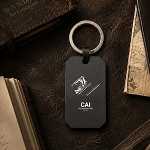 Könnte beinhalten: Ein rechteckiger Schlüsselanhänger aus schwarzem Metall mit einer weißen Grafik des Start- und Landebahnlayouts des Cairo International Airport, dem Text CAI und dem darunter gedruckten Schriftzug CAIRO INTERNATIONAL AIRPORT. Mit silbernem Metallring.