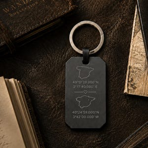 May include: A rectangular black metal keyring featuring two engraved outlines of Spain, each paired with specific geographic coordinates and separated by a small heart symbol. The keyring includes a silver-toned metal split ring at the top.