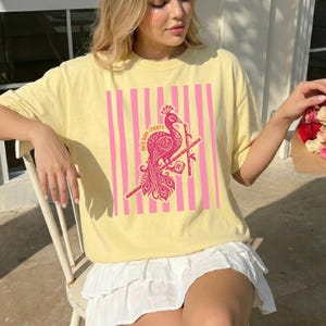 May include: A light yellow t-shirt featuring a pink peacock design with the text "bird ham cheers" on a pink striped background. The shirt is a casual style.