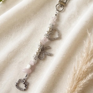 Puede incluir: Un llavero plateado con un cierre en forma de corazón y una cadena de dijes. La cadena presenta cuentas de piedras preciosas rosas, perlas de imitación, un corazón con pedrería, un lazo y un dije de Hello Kitty. El llavero está sobre un fondo de tela blanca.
