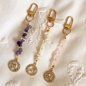 May include: Three keyrings with gold-coloured clasps and charms. Each keyring has a different gemstone bead strand: purple, white, and pink. The charms display zodiac symbols: Scorpio, Sagittarius, and Leo.