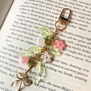 Puede incluir: Un llavero dorado con un dise&ntilde;o floral decorativo. El dije presenta flores rosas y blancas, hojas verdes y un lazo delicado. El llavero se muestra sobre el fondo de un libro abierto.